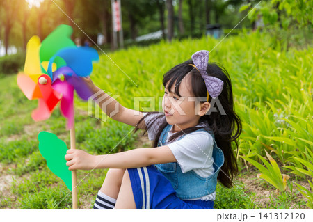 A cheerful girl sits comfortably in green foliage, looking at and holding a colorful toy pinwheel. 141312120