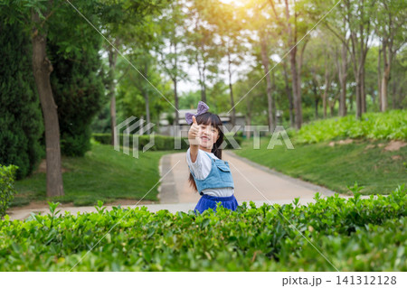 A joyful young girl with a bow stands on a park path, raising one arm up high with a bright smile. 141312128