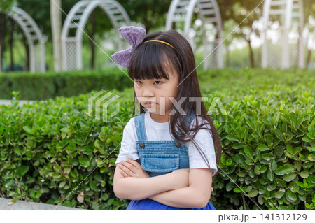 Against the backdrop of a lush garden, a young girl in denim overalls, arms crossed, looks angrily away, not at the camera. Against the backdrop of a lush garden, a young girl in denim overalls, arms crossed, looks angrily away, not at the camera. 141312129