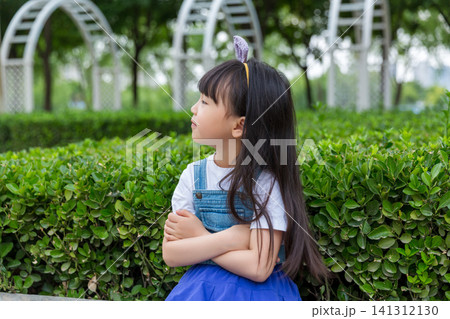 A thoughtful young girl in denim overalls with arms crossed looks away from the camera in a lush green garden setting. A thoughtful young girl in denim overalls with arms crossed looks away from the camera in a lush green garden setting. 141312130