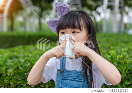 A young girl with a bow headband uses a white tissue to blow her nose while standing in a park with hedges. 141312132