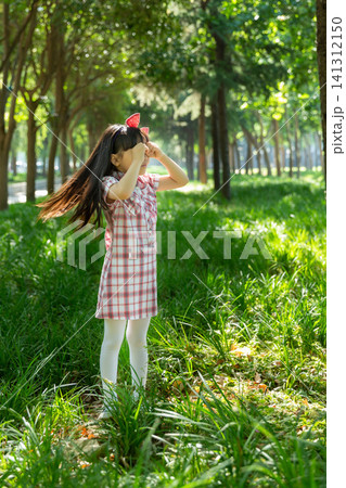 A joyful young girl in a plaid dress stands in a lush green grassy field, using both hands to make an eye-glass gesture. 141312150