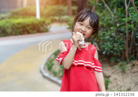 Cute young girl taking a big bite of a sandwich outdoors against a blurred green background. 141312156