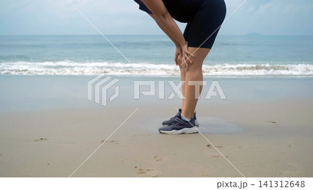 Young woman in black sportswear bending forward to catch her breath after running on the beach with sea waves visible behind her. Recovery moment concept Young woman in black sportswear bending forward to catch her breath after running on the beach with sea waves visible behind her. Recovery moment concept 141312648