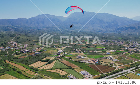 Horizontal aerial view of a paramotor paraglider flying high in the blue sky over the scenic green valley and majestic mountains of Crete island in Greece Horizontal aerial view of a paramotor paraglider flying high in the blue sky over the scenic green valley and majestic mountains of Crete island in Greece 141313390