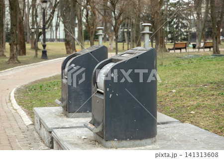A pair of modern metal waste containers installed on a concrete platform in a public city park, promoting urban cleanliness and efficient environmental management in a green space A pair of modern metal waste containers installed on a concrete platform in a public city park, promoting urban cleanliness and efficient environmental management in a green space 141313608