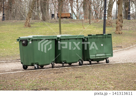 Three large green dumpsters sit on a paved path in a city park, showing an essential waste management system for keeping urban public spaces clean, orderly, and hygienic for all Three large green dumpsters sit on a paved path in a city park, showing an essential waste management system for keeping urban public spaces clean, orderly, and hygienic for all 141313611