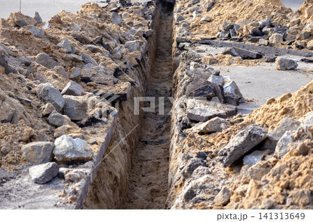 A long narrow trench dug in the ground for utility pipe installation, surrounded by piles of broken asphalt and soil at an urban construction site during road infrastructure repair A long narrow trench dug in the ground for utility pipe installation, surrounded by piles of broken asphalt and soil at an urban construction site during road infrastructure repair 141313649