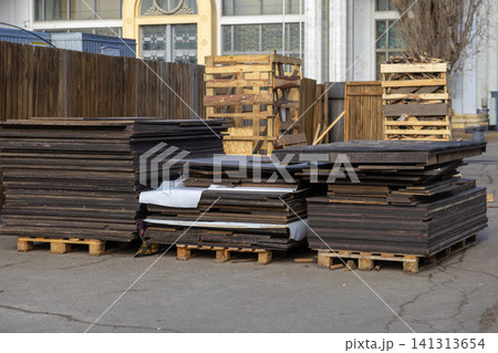 Stacked plywood boards on wooden pallets sit on an asphalt lot near a building exterior, representing industrial storage, construction materials management, and warehouse logistics 141313654