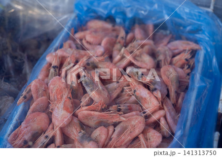 Pile of raw frozen pink shrimp inside a blue plastic container at a busy wholesale market, representing fresh seafood supply for commercial restaurant cooking and culinary industry Pile of raw frozen pink shrimp inside a blue plastic container at a busy wholesale market, representing fresh seafood supply for commercial restaurant cooking and culinary industry 141313750