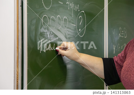 A teacher writes complex math exercises with white chalk on a green classroom blackboard to represent primary school education, academic instruction, and learning in a school 141313863