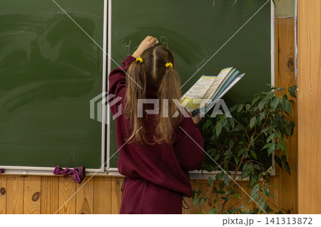 A young schoolchild writes on a green chalkboard while holding a textbook in a classroom, representing primary education, academic growth, learning, concentration and development A young schoolchild writes on a green chalkboard while holding a textbook in a classroom, representing primary education, academic growth, learning, concentration and development 141313872