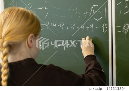 A young schoolchild writes complex math equations on a green classroom blackboard using white chalk while engaged in focused learning during an elementary school mathematics lesson 141313894