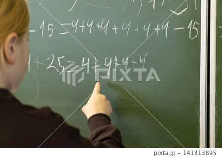 A young schoolchild points at complex mathematical equations on a green chalkboard in a primary classroom, highlighting interactive learning and the process of academic solving A young schoolchild points at complex mathematical equations on a green chalkboard in a primary classroom, highlighting interactive learning and the process of academic solving 141313895