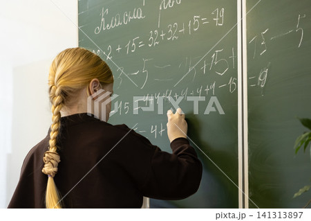 A blonde schoolchild writes complex math equations on a green chalkboard in an elementary classroom, representing academic learning, mental development, and focus in early education 141313897