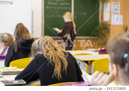 A young schoolchild stands at a chalkboard reading a book in a primary school classroom while peers work at desks, capturing an academic environment of learning and participation A young schoolchild stands at a chalkboard reading a book in a primary school classroom while peers work at desks, capturing an academic environment of learning and participation 141313900
