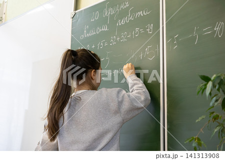 A young schoolchild writes complex math equations with white chalk on a green chalkboard in a primary classroom to represent academic learning and mental development for students A young schoolchild writes complex math equations with white chalk on a green chalkboard in a primary classroom to represent academic learning and mental development for students 141313908