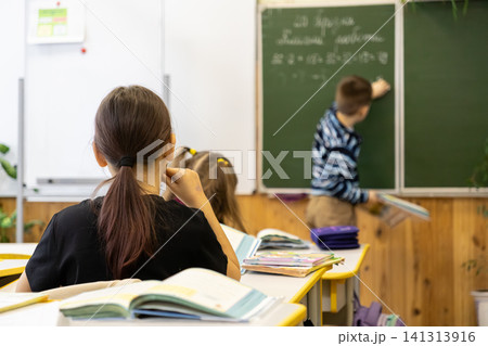 Young students sit at wooden desks in an elementary school classroom while a classmate writes on a green chalkboard to capture the focused atmosphere of primary education lessons Young students sit at wooden desks in an elementary school classroom while a classmate writes on a green chalkboard to capture the focused atmosphere of primary education lessons 141313916