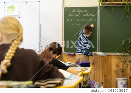 A young schoolchild writes complex math equations on a green chalkboard in an elementary classroom while peers study at desks, highlighting active participation in primary school A young schoolchild writes complex math equations on a green chalkboard in an elementary classroom while peers study at desks, highlighting active participation in primary school 141313917