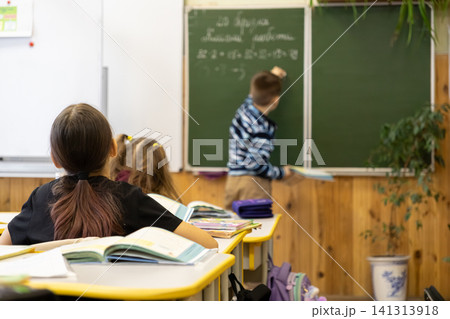Primary school students sit at desks watching a classmate solve a math problem on the green chalkboard, illustrating a focused academic learning environment in a modern classroom Primary school students sit at desks watching a classmate solve a math problem on the green chalkboard, illustrating a focused academic learning environment in a modern classroom 141313918