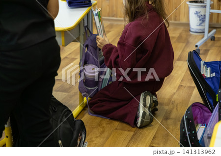 A young schoolchild kneels by a wooden desk to pack a purple backpack in an elementary classroom, capturing daily academic preparation and student organization routines for kids A young schoolchild kneels by a wooden desk to pack a purple backpack in an elementary classroom, capturing daily academic preparation and student organization routines for kids 141313962