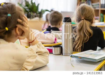 Group of young schoolchildren sitting at wooden desks in a bright elementary classroom with metal thermoses and books, representing focused learning and daily academic education 141313963