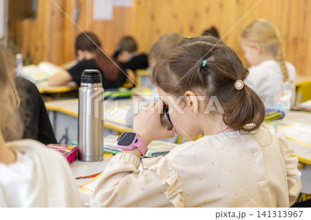 Schoolchild drinking water from a metal bottle during a primary school lesson, sitting at a desk surrounded by other students in a bright classroom setting for academic development 141313967
