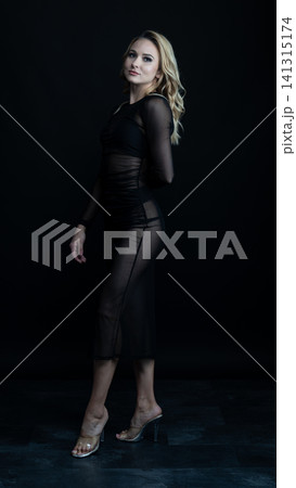 Young blonde dancer wearing transparent black dress and high heels posing in studio with black background 141315174