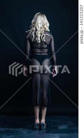 Young blonde dancer wearing transparent black dress and high heels posing in studio with black background 141315207