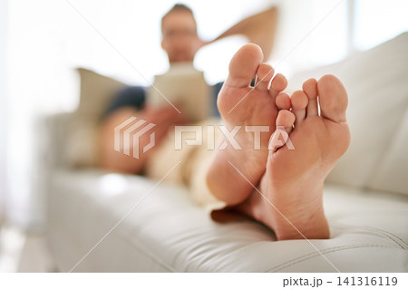 Feet, relax and man on sofa in living room reading a book, novel or story for knowledge at home. Rest, chill and closeup to male person foot laying on couch in lounge of modern apartment on weekend. Feet, relax and man on sofa in living room reading a book, novel or story for knowledge at home. Rest, chill and closeup to male person foot laying on couch in lounge of modern apartment on weekend. 141316119