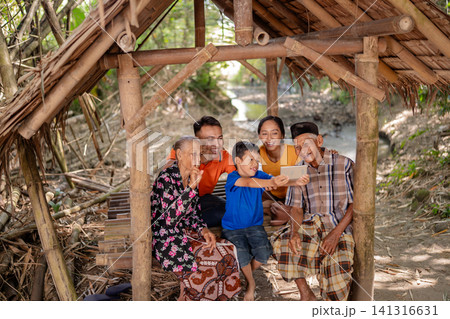 Multigenerational family smiling together under rustic bamboo shelter while young boy holds smartphone for selfie, with grandparents, parents, and child gathered outdoors in warm daylight Multigenerational family smiling together under rustic bamboo shelter while young boy holds smartphone for selfie, with grandparents, parents, and child gathered outdoors in warm daylight 141316631