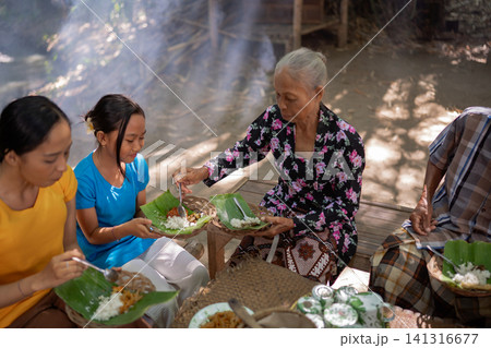 Three generations sharing traditional meal outdoors on woven tableware with rice, banana leaves, smoke, and warm sunlight in rustic village setting during family gathering Three generations sharing traditional meal outdoors on woven tableware with rice, banana leaves, smoke, and warm sunlight in rustic village setting during family gathering 141316677