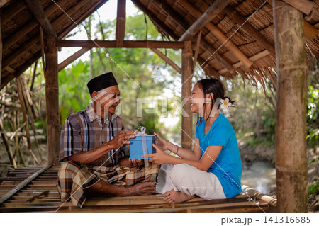 Elderly man and young woman smiling while exchanging blue gift box inside rustic bamboo shelter with natural daylight, warm intergenerational scene, cultural setting, and relaxed outdoor atmosphere Elderly man and young woman smiling while exchanging blue gift box inside rustic bamboo shelter with natural daylight, warm intergenerational scene, cultural setting, and relaxed outdoor atmosphere 141316685