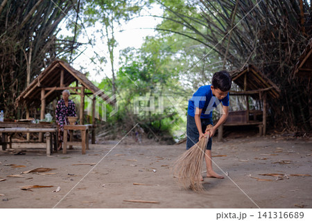 Young boy sweeping dry ground beside rustic bamboo huts in rural village yard with elderly woman nearby, framed by tall arching trees and soft daylight in calm outdoor setting 141316689