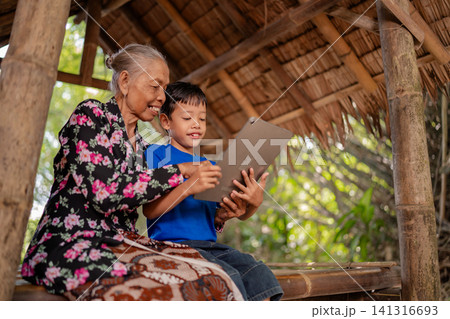 Elderly woman and young boy sharing tablet screen inside rustic bamboo shelter, smiling together in warm daylight with family learning, intergenerational bonding, and rural lifestyle atmosphere Elderly woman and young boy sharing tablet screen inside rustic bamboo shelter, smiling together in warm daylight with family learning, intergenerational bonding, and rural lifestyle atmosphere 141316693
