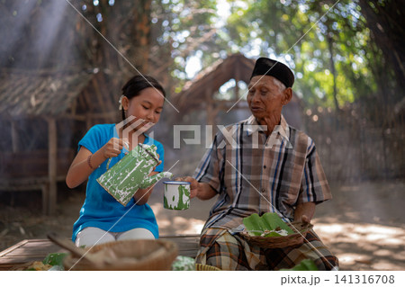 Young girl pours tea from enamel pot for elderly man during outdoor meal in shaded village setting, with traditional clothing, woven trays, soft sunlight, and calm family atmosphere 141316708