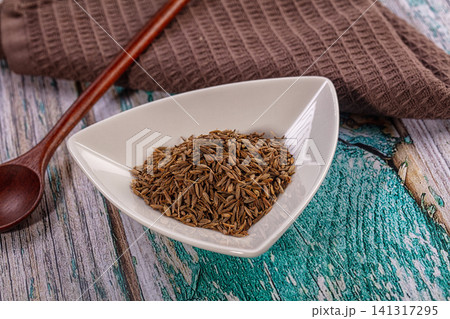 Cumin seeds in white ceramic bowl on rustic wooden table with brown cloth and wooden spoon 141317295