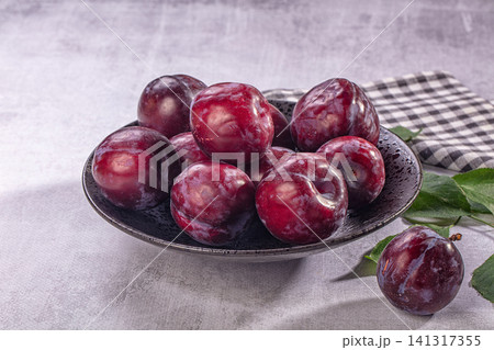 Fresh ripe plums in dark ceramic bowl on textured gray surface with checkered cloth and green leaves 141317355