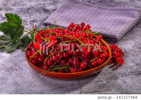 Fresh red currants in wooden bowl on stone surface with green leaves and purple cloth 141317368