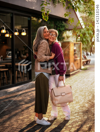 Warm Street Reunion of Two Stylish Mature Women Sharing a Full Embrace Outside a Cozy Urban Cafe in Golden Afternoon Light Warm Street Reunion of Two Stylish Mature Women Sharing a Full Embrace Outside a Cozy Urban Cafe in Golden Afternoon Light 141318146