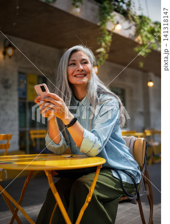 Radiant Mature Woman Holding Smartphone and Smiling Away from Camera at a Bright Yellow Outdoor Cafe Table in Warm Ambient Light and Trailing Greenery Radiant Mature Woman Holding Smartphone and Smiling Away from Camera at a Bright Yellow Outdoor Cafe Table in Warm Ambient Light and Trailing Greenery 141318147