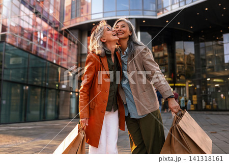 Joyful Mature Women Friends Sharing a Playful Cheek Kiss Outside a Modern Glass Shopping Center Holding Kraft Bags in Earth Tone Outfits Joyful Mature Women Friends Sharing a Playful Cheek Kiss Outside a Modern Glass Shopping Center Holding Kraft Bags in Earth Tone Outfits 141318161