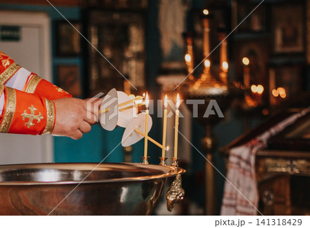 Orthodox priest lighting baptism candles during a Christian church ceremony. Close-up of sacred ritual with holy water, candles and traditional liturgical vestments, symbolizing faith, spirituality 141318429