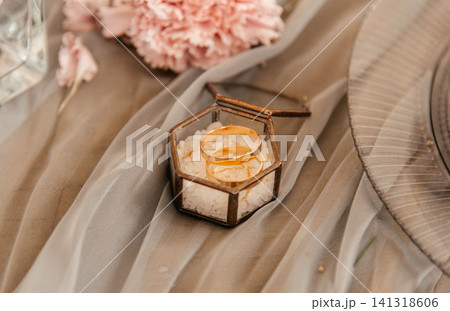 Elegant gold wedding rings displayed in a geometric glass ring box filled with decorative salt. Romantic wedding table decor with soft fabric, pink carnation and stylish details. Close-up symbolizing Elegant gold wedding rings displayed in a geometric glass ring box filled with decorative salt. Romantic wedding table decor with soft fabric, pink carnation and stylish details. Close-up symbolizing 141318606