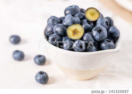 Fresh blueberries are piled into a small rustic ceramic bowl, with a few sliced berries revealing their green centers on a white fabric surface 141320087