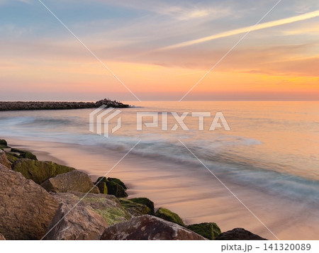 Serene sunset over the Atlantic at Praia do Furadouro in Ovar, Portugal, with glowing orange skies reflecting on the wet sand and mossy rocks Serene sunset over the Atlantic at Praia do Furadouro in Ovar, Portugal, with glowing orange skies reflecting on the wet sand and mossy rocks 141320089