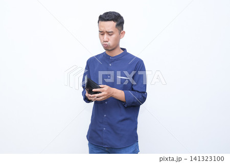 Young man holds wallet, touches forehead in stress. He wears blue shirt, shows frustration or disappointment. Blank background emphasizes emotional moment. Ideal for financial struggles 141323100