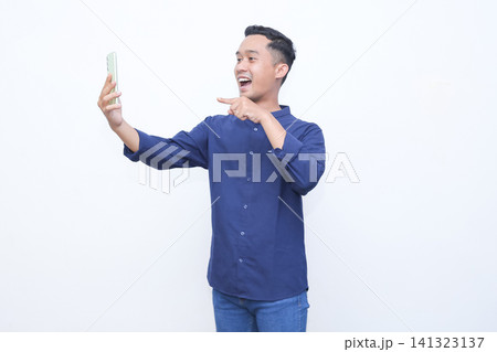 Young man laughs joyfully while holding phone. He points toward camera with cheerful expression. Bright background highlights his lively mood. Ideal for social media, influencer, or lifestyle content 141323137