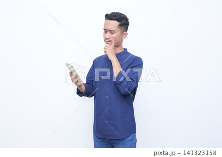 Young man in blue shirt holds phone, ponders thoughtfully. Hand on chin, eyes upward, shows deep reflection. White background isolates subject, emphasizes expression. Ideal for business, Young man in blue shirt holds phone, ponders thoughtfully. Hand on chin, eyes upward, shows deep reflection. White background isolates subject, emphasizes expression. Ideal for business, 141323158