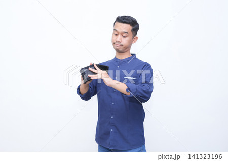 Young man holds wallet, touches forehead in stress. He wears blue shirt, shows frustration or disappointment. Blank background emphasizes emotional moment. Ideal for financial struggles 141323196